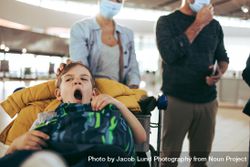 Boy yawning while sitting on luggage trolley at airport with parents in background bErmN0