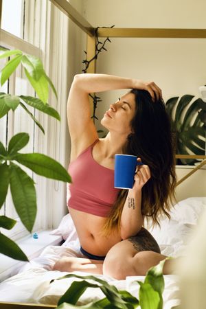 Woman holding a blue mug sitting on bed near window