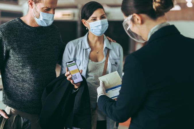 Male and female traveler in face masks at check-in counter displaying vaccine passport to staff