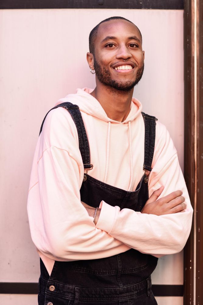 Smiling male in denim overalls and earrings with arms crossed against a pink wall