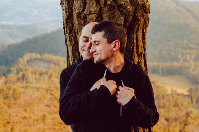 Smiling men hugging near tree in forest on hill