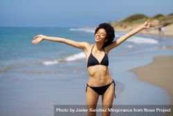 Woman in bikini standing on beach with outstretched arms and eyes closed bD1dr0