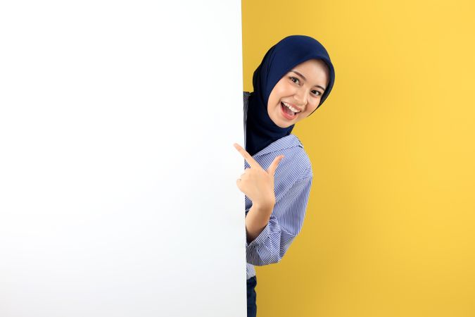 A Joyful Asian Woman in Blue Navy Hijab and Striped Shirt Excitedly Points at a Blank Board