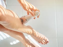 Young white woman depilates her leg with an electric epilator device in the bathroom.
