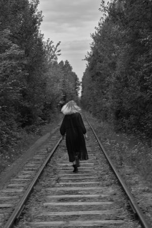 Back view of woman in dark coat walking on train rail in grayscale