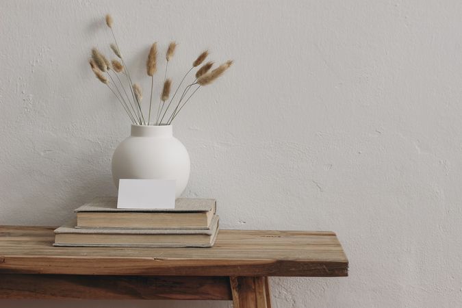 Blank business card mockup on old books. Modern round ceramic vase with dry bunny tails, Lagurus ovatus grass on wooden table, bench. Scandinavian interior, home office.