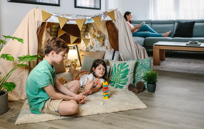 Little girl catching piece of stacking game while play with brother and mother reading book on sofa