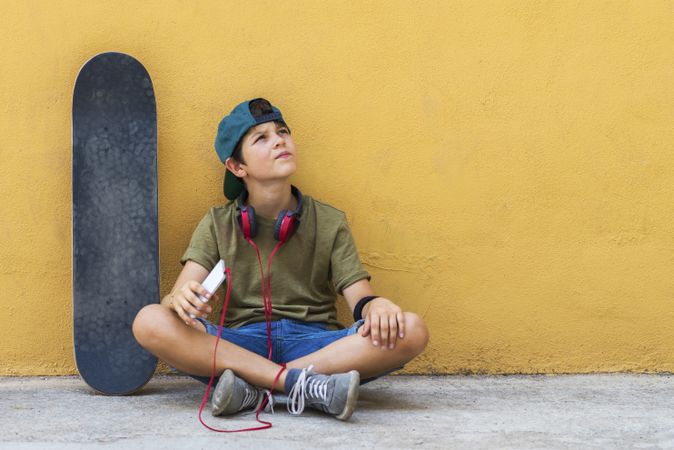 Front view of a teenage boy sitting on ground leaning on a yellow wall while holding a mobile phone