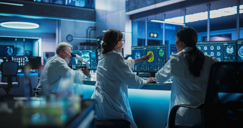 Male Doctor Using Microscope And His Female Colleagues Looking At MRI Brain Scans On Desktop Computers.