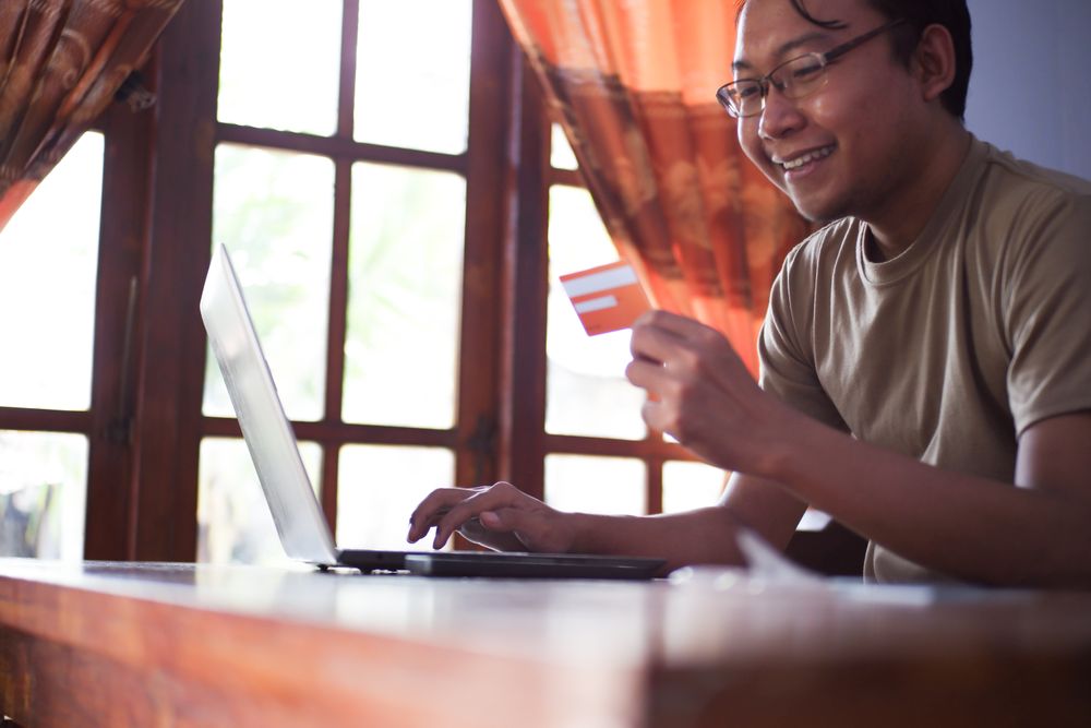 Asian Male with shirt doing online transaction in living room with credit card smartphone and laptop