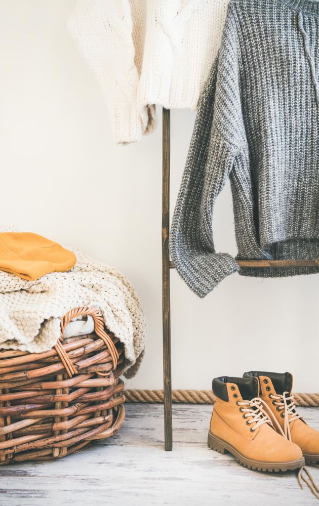 Woolen sweaters, grey scarf, winter boots on modern garment rack, with thatched basket, close up