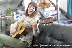 Female on sofa playing acoustic guitar and singing at home in living room 4AkJ80