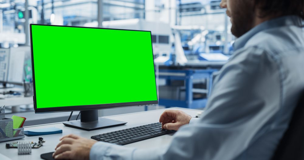 Young Industrial Software Engineer Working on a Desktop Computer with Green Screen Mock-Up Display. Modern Factory with High-Tech Machinery and Robotic Arms in the Assembly Hall