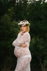 Pregnant woman with closed eyes in white lace dress touching chin while standing in forest