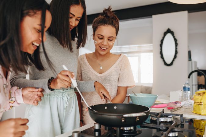 Young women frying food in pan standing with her friends in kitchen