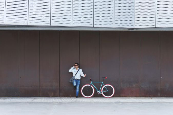 Male in sunglasses standing with red and green bicycle in front of wall and talking on phone
