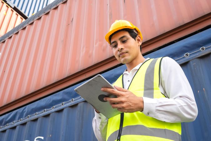 Man in hard hat standing beside shipping container with tablet