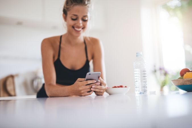 Happy young woman standing in kitchen reading messages on her cell phone