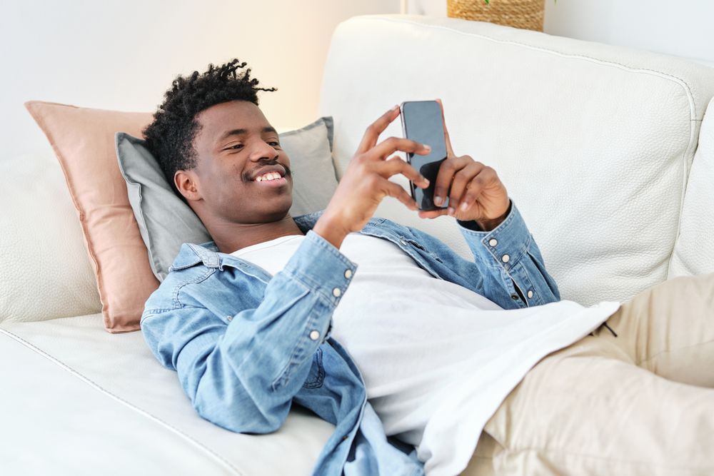 Happy Black man lying on sofa and using smartphone