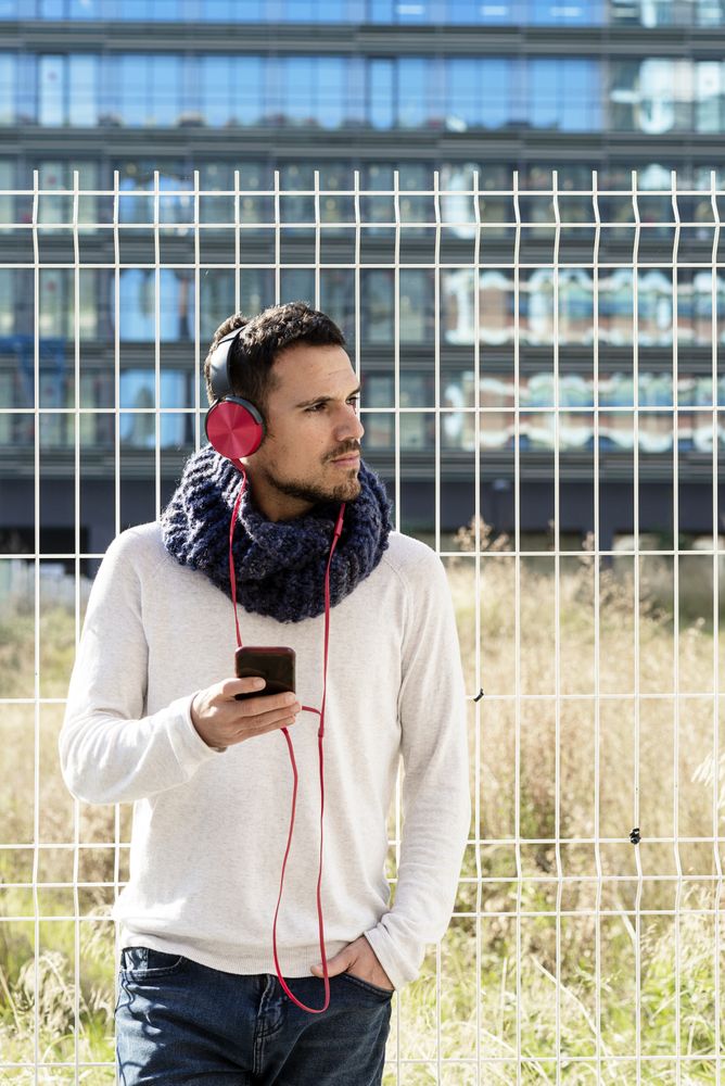 Man walking in scarf leaning back on metallic fence and looking up from using smartphone
