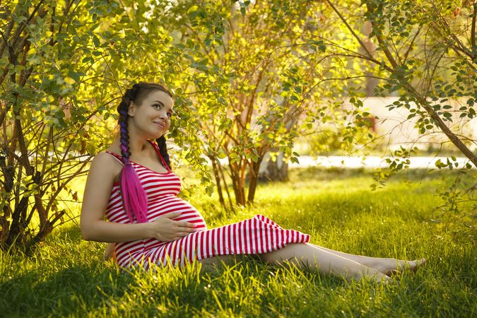 Pregnant female in red striped dress sitting under trees on the grass