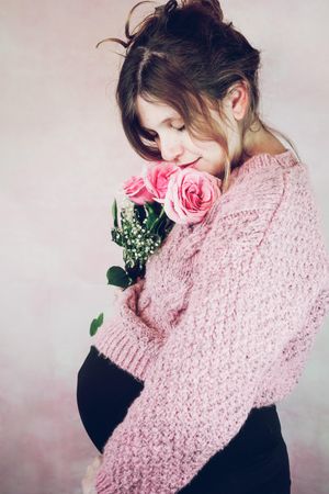 Side view of pregnant woman in pink knit sweater holding rose bouquet against pink background