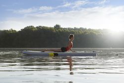 Side view of woman stretching back on paddleboard on fresh lake in the morning surrounded by trees
