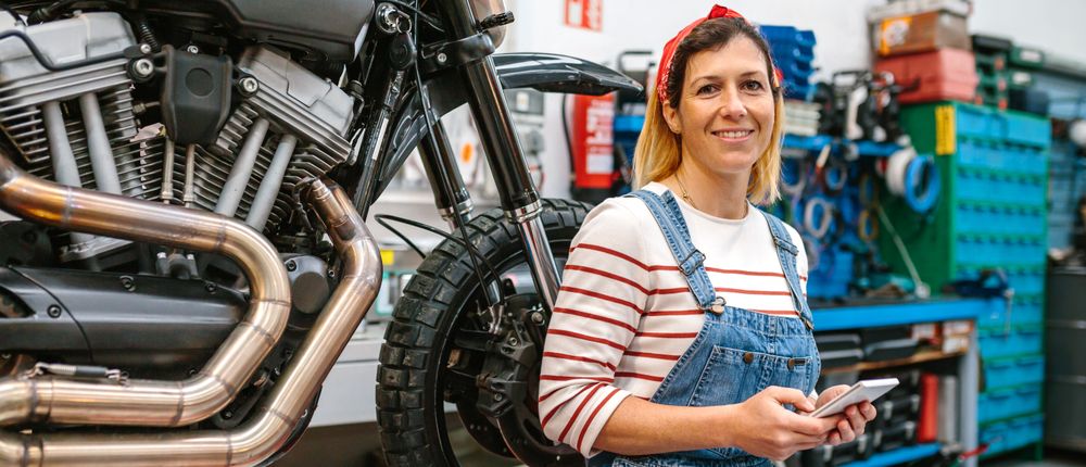Mechanic woman holding phone sitting over platform with motorcycle on factory