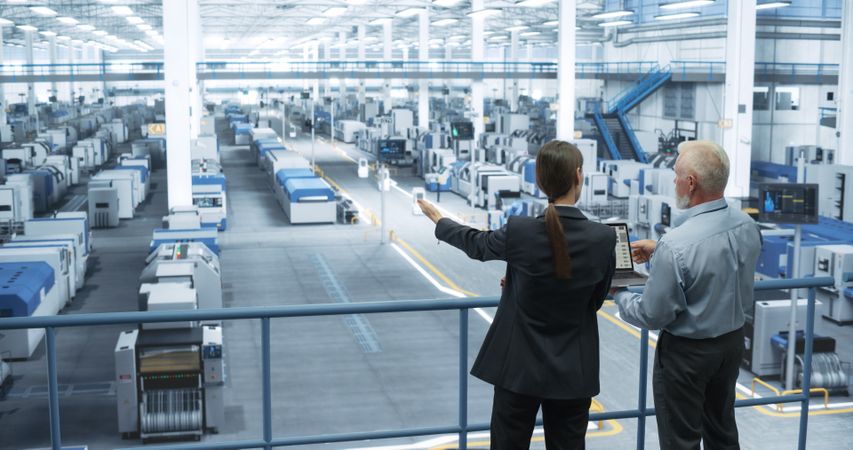 Male Engineer and Female Scientist Standing with Their Back, Using Laptop Computer and Talking in a Factory Facility with Equipment Producing Modern Technical Devices for Different Industries
