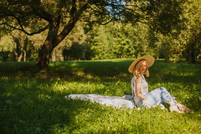Woman with sunhat sitting on light textile on green grass field near trees