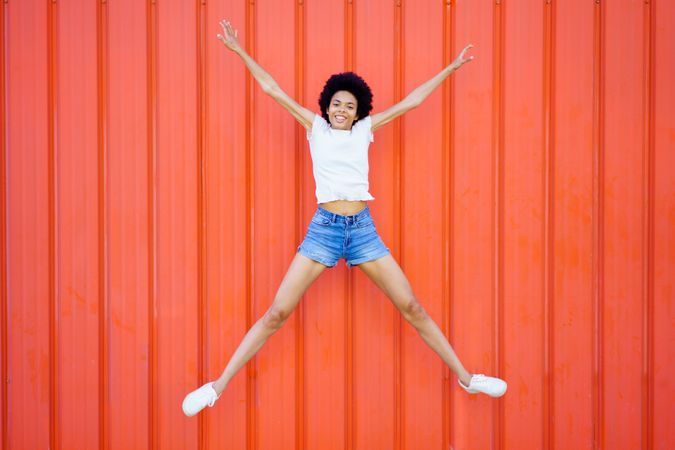 Woman in shorts and t-shirt jumping for joy against red wall