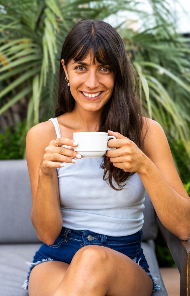 A woman is lookintg at camera while sitting on a couch and holding a white cup. She is smiling and she is enjoying her coffee