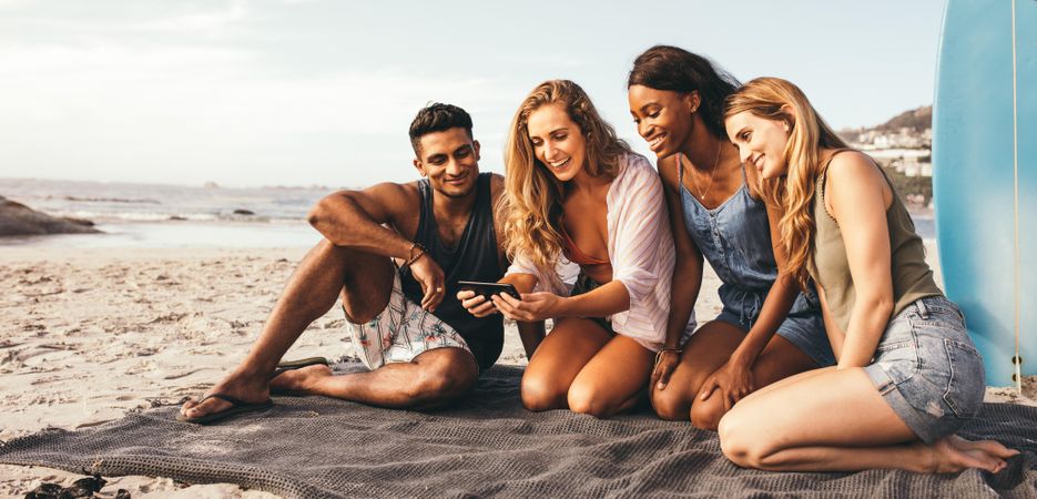 Smiling woman taking selfie with her friends sitting on a beach with a surfboard in the background
