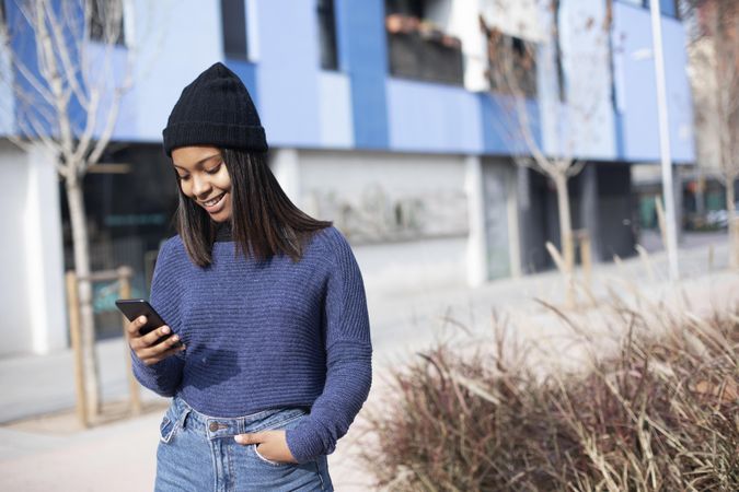 Smiling female in wool hat and sweater checking phone outside blue building