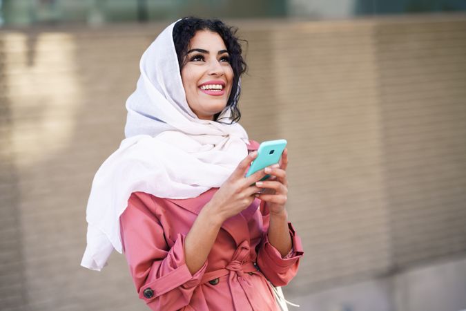 Happy female in headscarf and pink trench coat sitting outside with her phone