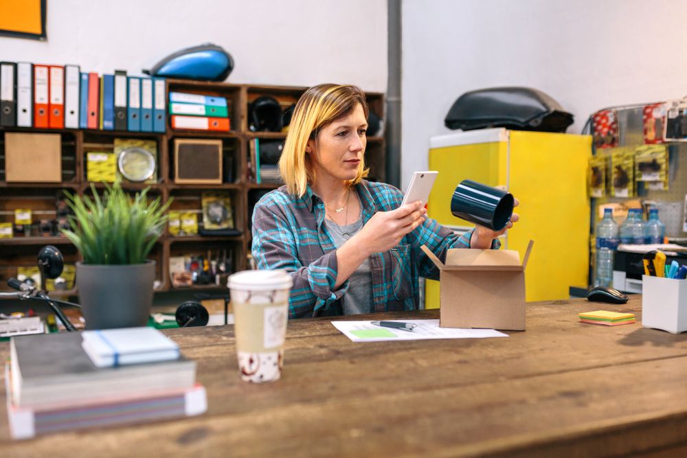 Young female entrepreneur taking photo with cell of a blue mug for client of her online store