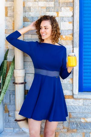 Happy woman in blue dress touching hair while holding juice outside of home