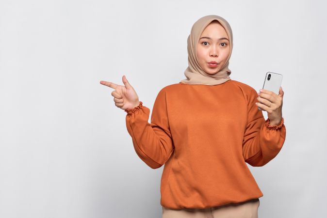 Surprised Muslim woman holding smart phone and pointing to her side