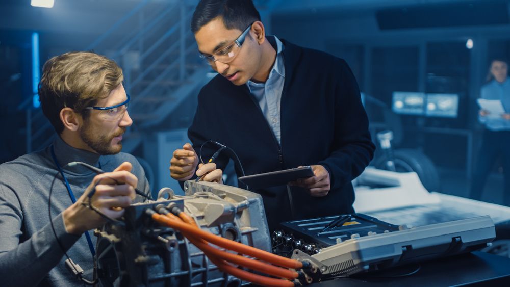 Two Professional Automotive Engineers with a Tablet Computer and Inspection Tools are Having a Conversation While Testing an Electric Engine in a High Tech Laboratory with a Concept Car Chassis.