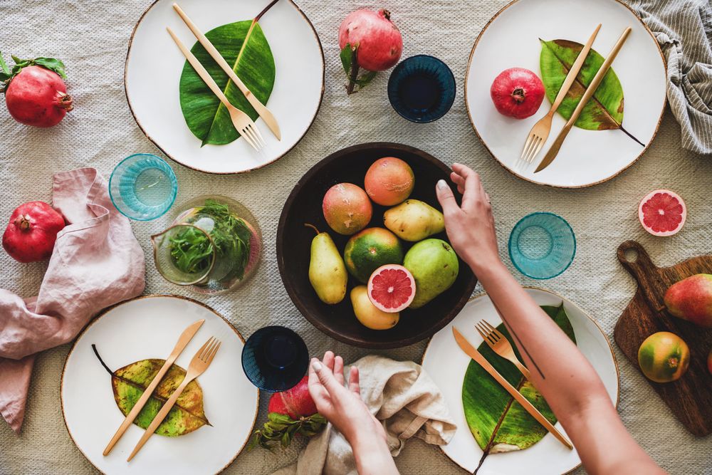 Fresh table setting with leaves on bright plates, glassware, with person reaching for fruit bowl