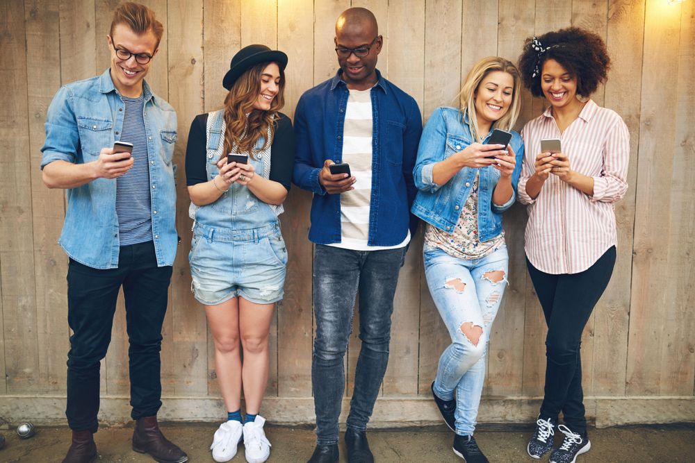 Multi-ethnic group of friends standing against a wooden fence sharing content on their phones