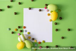 Easter eggs and chocolate pieces on green background with paper card square 0Vg3k5