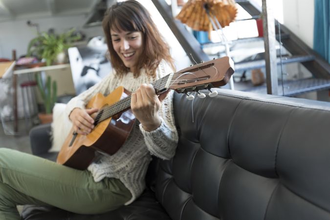Laughing female on sofa enjoying playing music of her acoustic guitar in living room