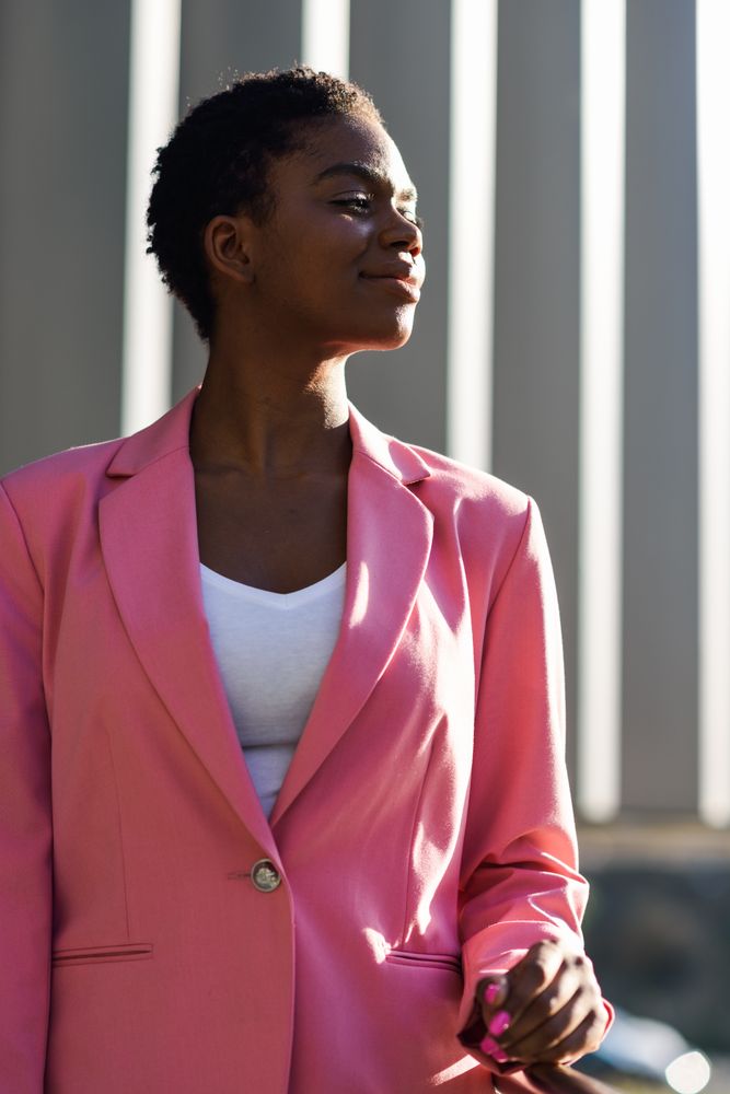 Female in suit with pink jacket outside office with striped shadows looking to her side, vertical
