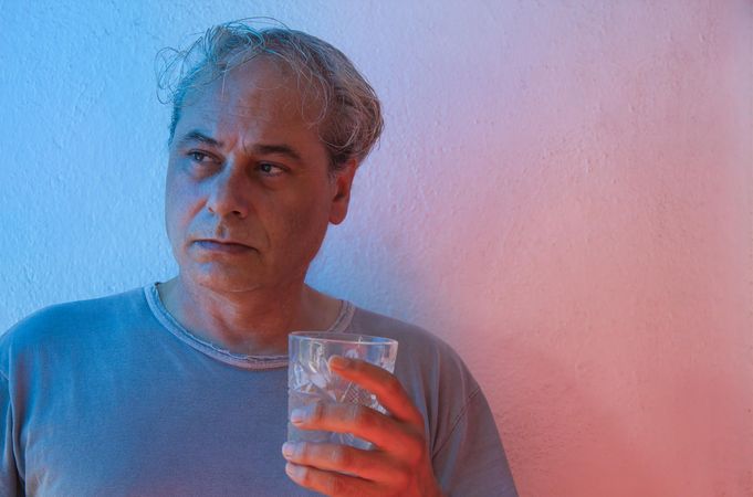 Portrait of serious middle aged man in gray shirt holding a glass of water against light background in UV lit studio