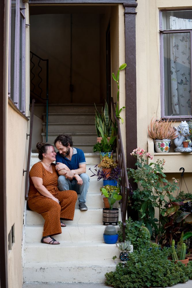 Happy couple smiling and looking at each other sitting next to houseplants on apartment stairs