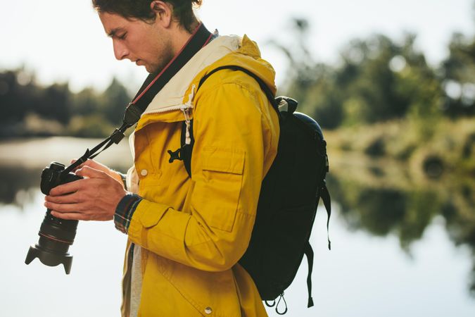 Close up of a man standing near a lake wearing a backpack looking at his dslr camera
