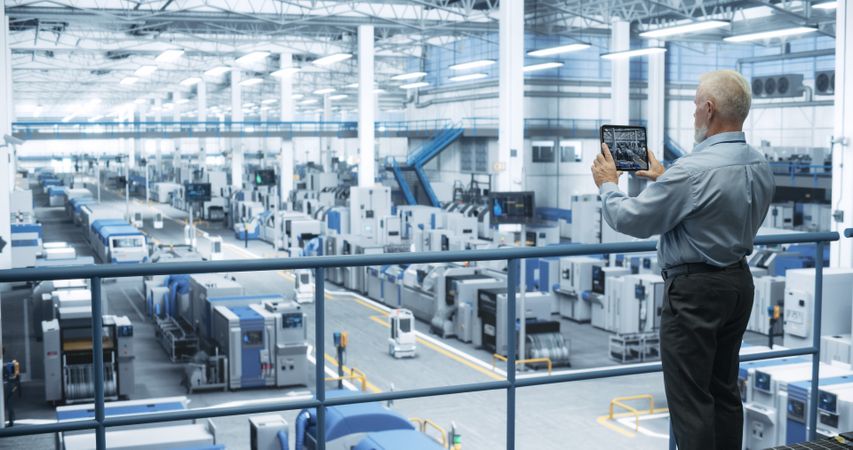 Mature Engineer Standing on a Platform, Using Tablet Computer at an Electronics Factory. Machines are Undergoing Maintenance, Specialist Monitoring the Progress Through Online Software
