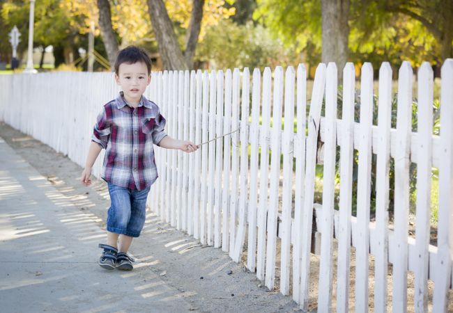 Young Biracial Boy Walking with Stick Along White Fence