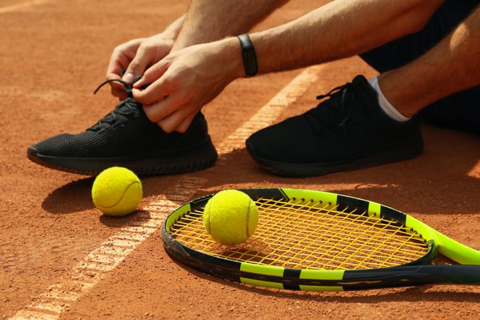 Man ties his shoelaces on clay court with racket and balls